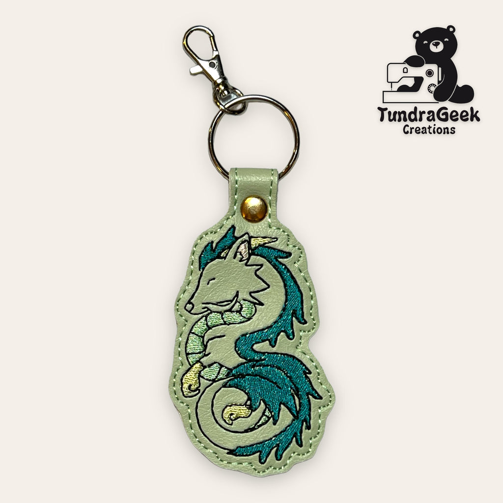 A keychain with an embroidered turquoise dragon design, attached to a metal keyring and lobster clasp. The TundraGeek Creations logo appears in the top right corner.