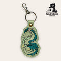 A keychain with an embroidered turquoise dragon design, attached to a metal keyring and lobster clasp. The TundraGeek Creations logo appears in the top right corner.