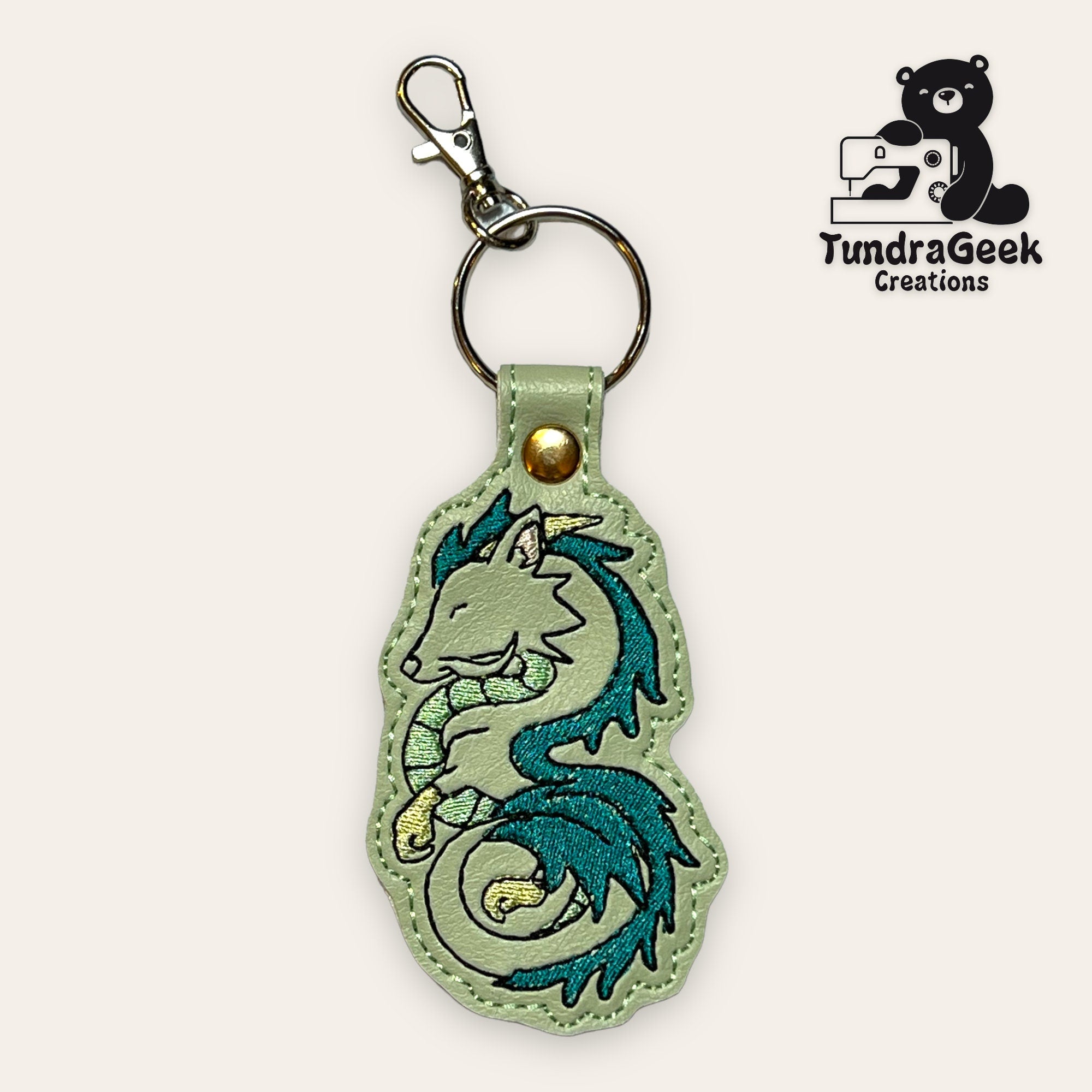 A keychain with an embroidered turquoise dragon design, attached to a metal keyring and lobster clasp. The TundraGeek Creations logo appears in the top right corner.