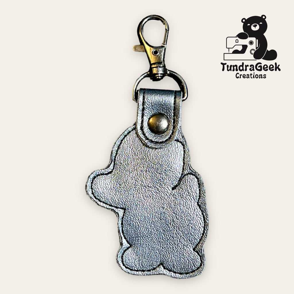 Silver bear-shaped leather keychain with metal clasp, displayed on a light background. TundraGeek Creations logo with a bear and sewing machine is in the top right corner.