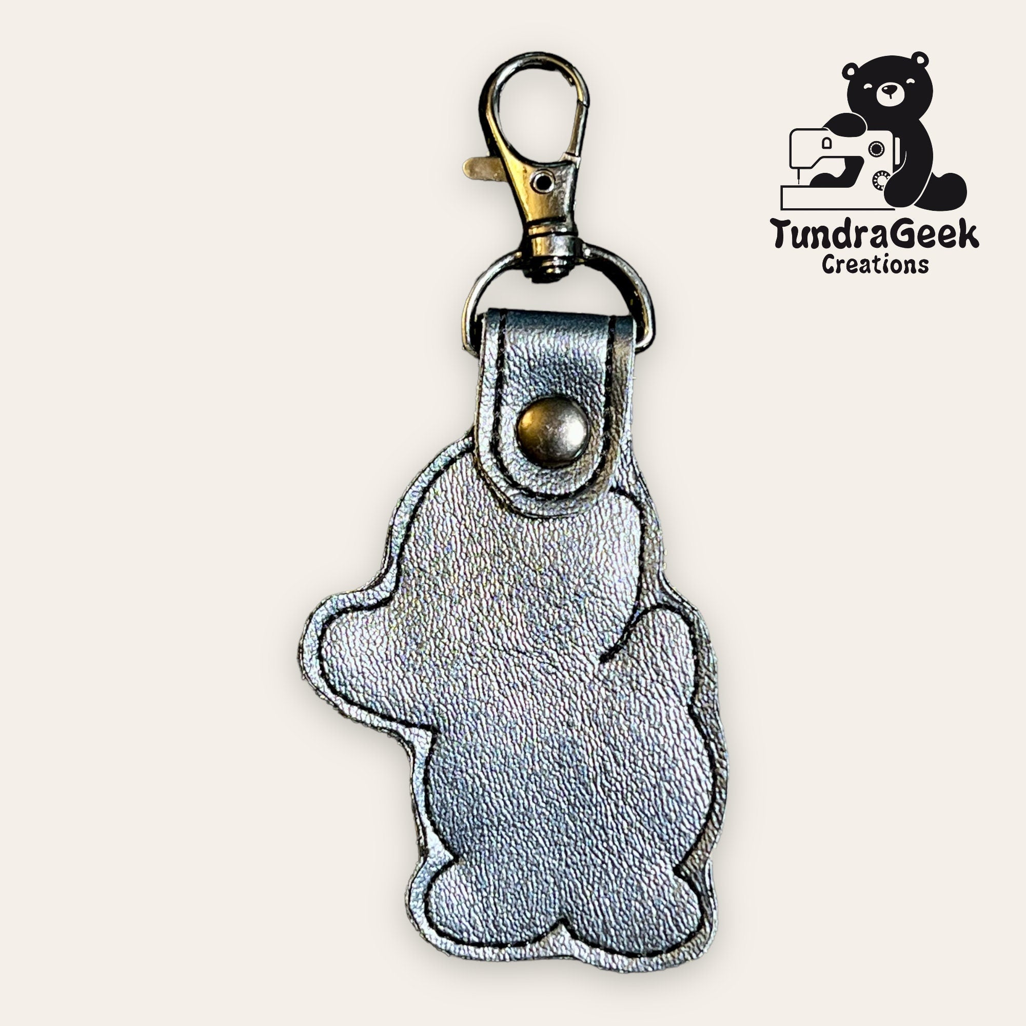 Silver bear-shaped leather keychain with metal clasp, displayed on a light background. TundraGeek Creations logo with a bear and sewing machine is in the top right corner.