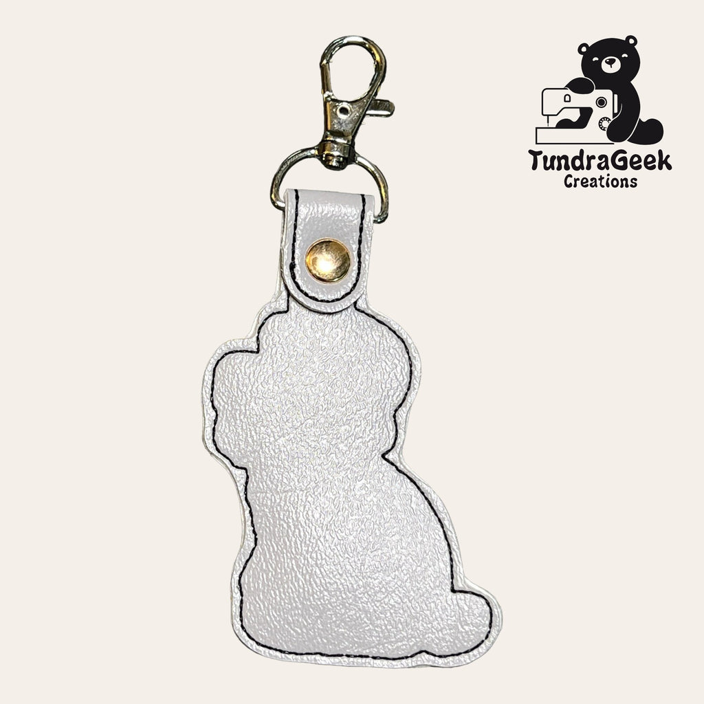 A white faux leather keychain shaped like a sitting dog with a metal clasp, next to the TundraGeek Creations logo featuring a bear and a sewing machine.