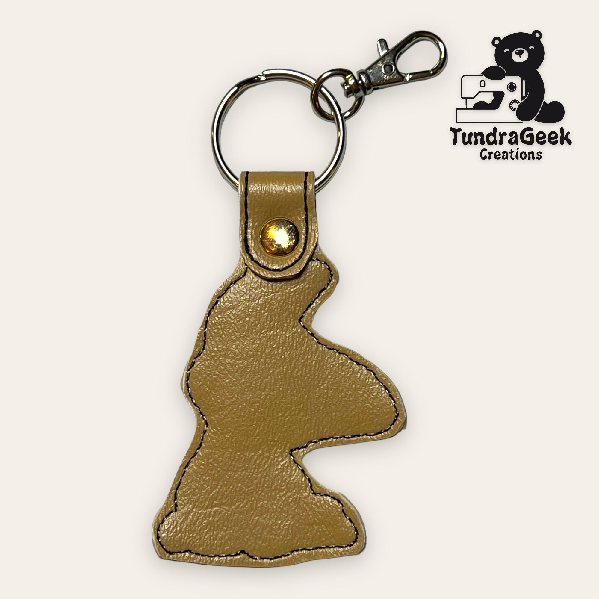 A tan leather keychain shaped like a cartoon dog head, attached to a metal keyring and clasp; TundraGeek Creations logo in the corner.