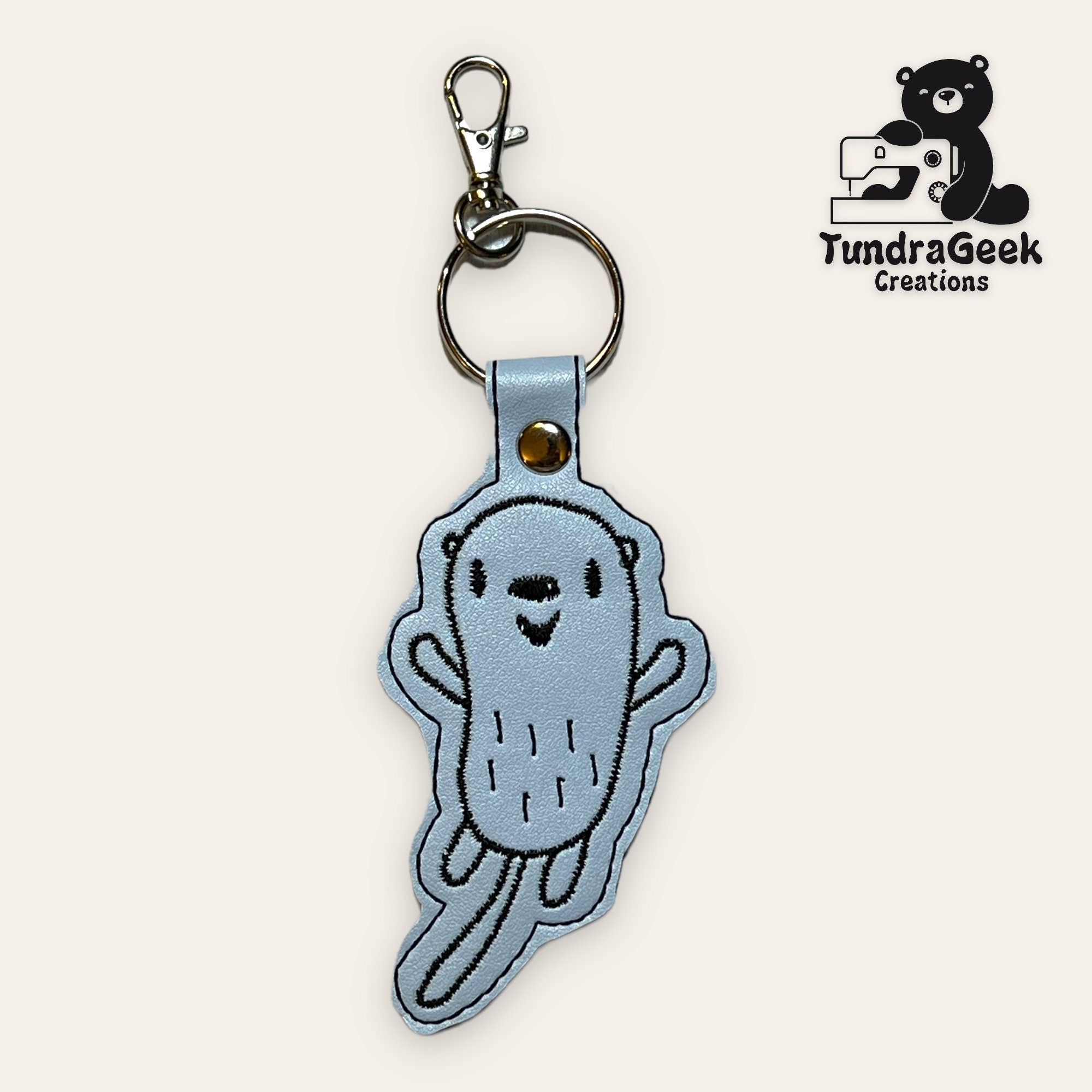 Light blue embroidered keychain featuring a cartoon otter with a smiling face, attached to a metal keyring and clasp. TundraGeek Creations logo is visible in the top right corner.