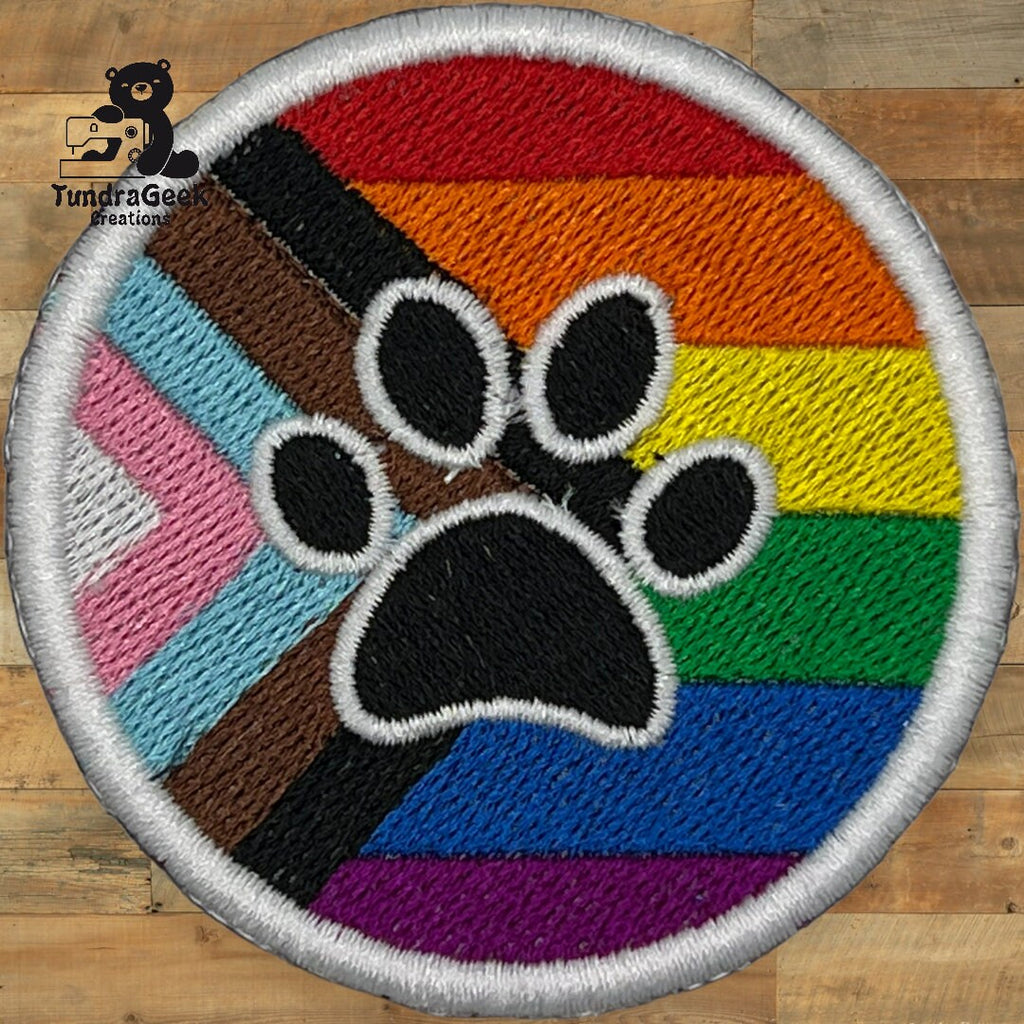 Round embroidered patch featuring a black paw print overlaid on a Progress Pride flag background, placed on a wooden surface.