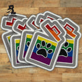 Embroidered patches featuring a paw print overlaid on a rainbow flag with a red heart above, arranged in a stack on a wooden surface.
