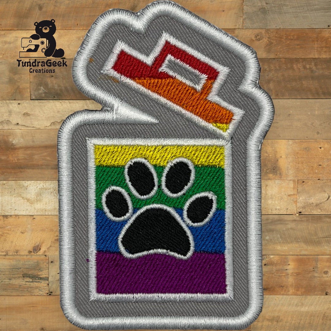 An embroidered patch shaped like an open lighter features a black paw print on a rainbow-striped background, displayed on a wooden surface.