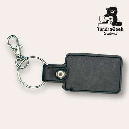 Black rectangular keychain with a metal key ring and clip, displayed on a light background next to the TundraGeek Creations logo featuring a sewing bear.