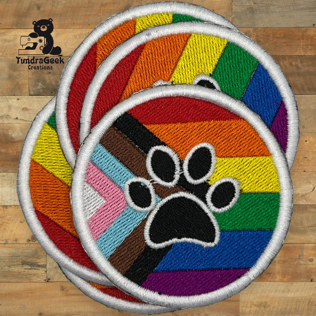 Three embroidered round patches featuring a paw print overlaid on the Progress Pride flag, displayed on a wooden surface.