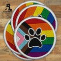 Three embroidered round patches featuring a paw print overlaid on the Progress Pride flag, displayed on a wooden surface.
