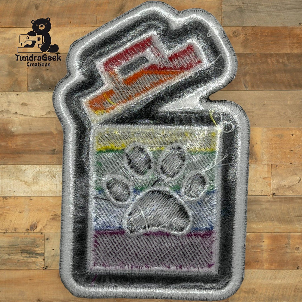 An embroidered patch shaped like a lighter with a paw print and rainbow stripes, displayed on a wooden background. TundraGeek Creations logo in the top left corner.