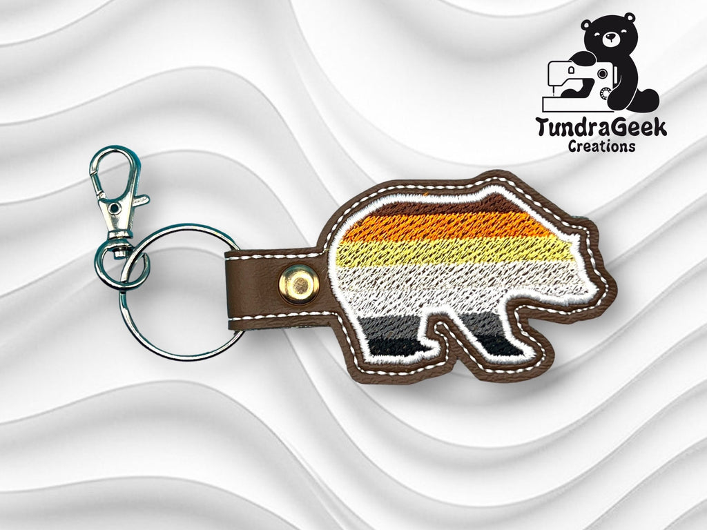 Embroidered bear keychain with brown, orange, yellow, and white stripes on a wavy white background, next to TundraGeek Creations logo.