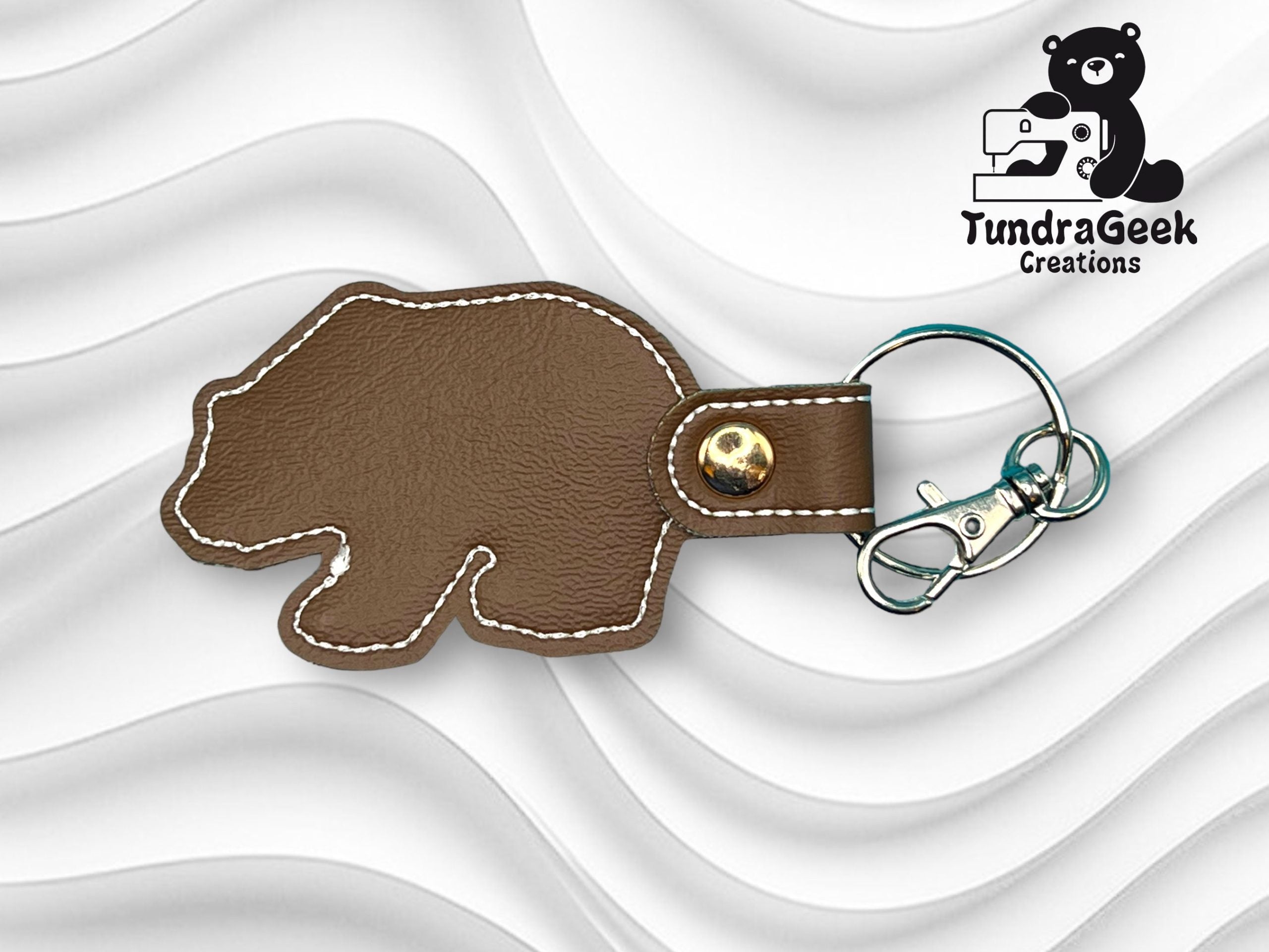 Brown bear-shaped leather keychain with white stitching and metal hardware on a wavy white background. TundraGeek Creations logo is in the top right corner.