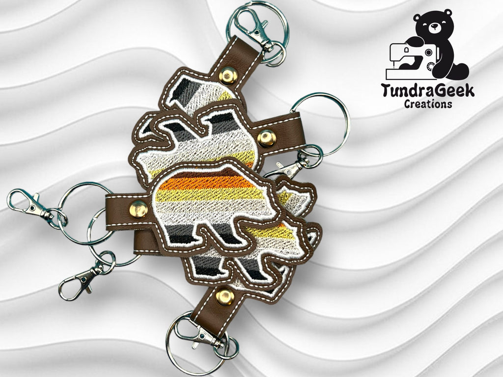 Three bear-shaped keychains with brown, yellow, orange, white, and black stripes on a wavy white background. Logo for TundraGeek Creations appears in the top right corner.