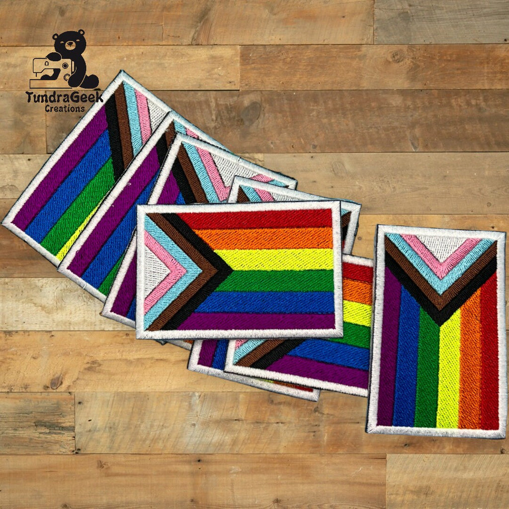 Five embroidered Pride flag patches, including Progress Pride designs, arranged overlapping on a wooden surface with a TundraGeek Creations logo in the corner.