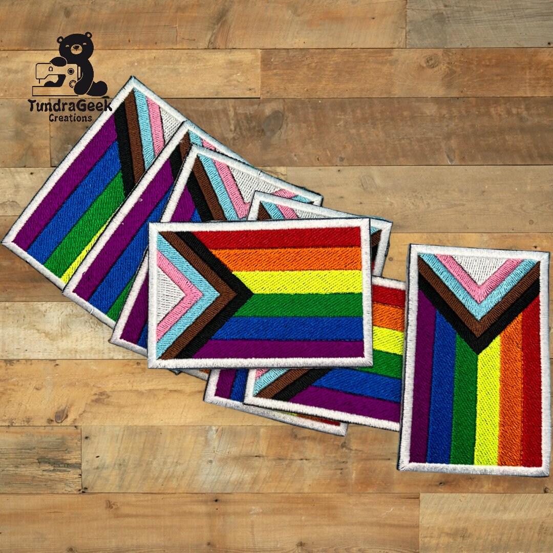 Five embroidered Pride flag patches, including Progress Pride designs, arranged overlapping on a wooden surface with a TundraGeek Creations logo in the corner.