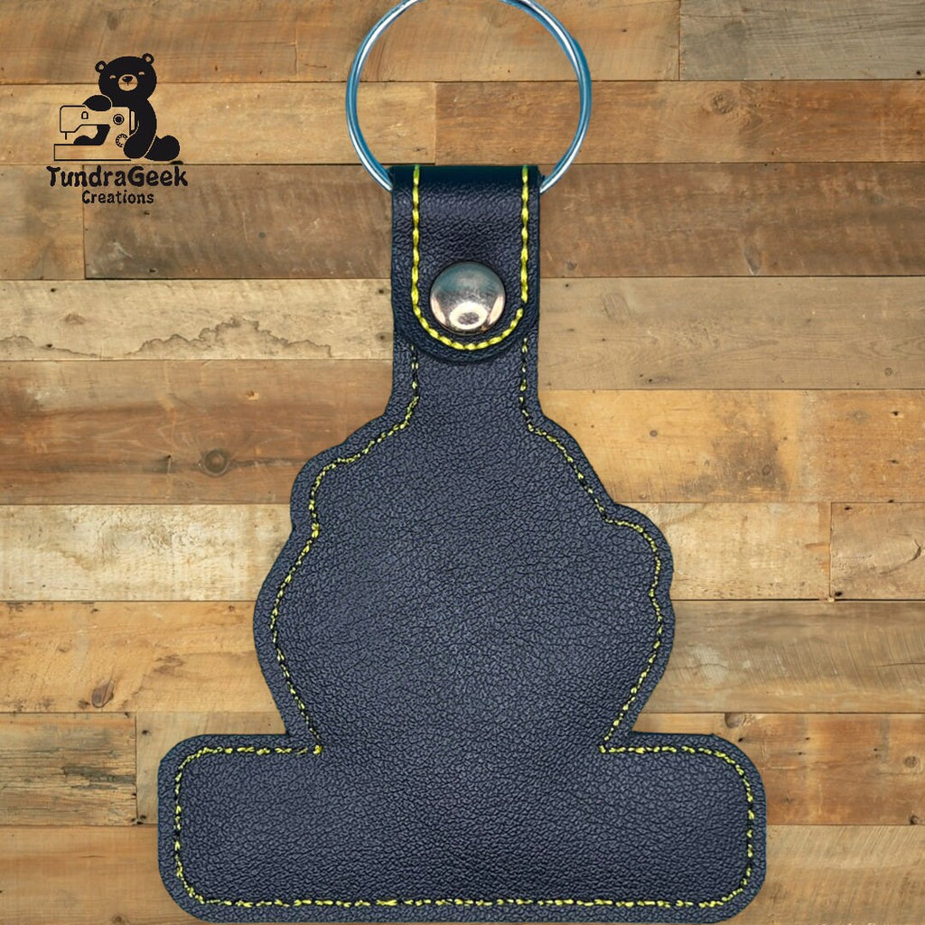 Black leather keychain with yellow stitching and metal ring, displayed on a wooden surface. TundraGeek Creations logo in the top left corner.