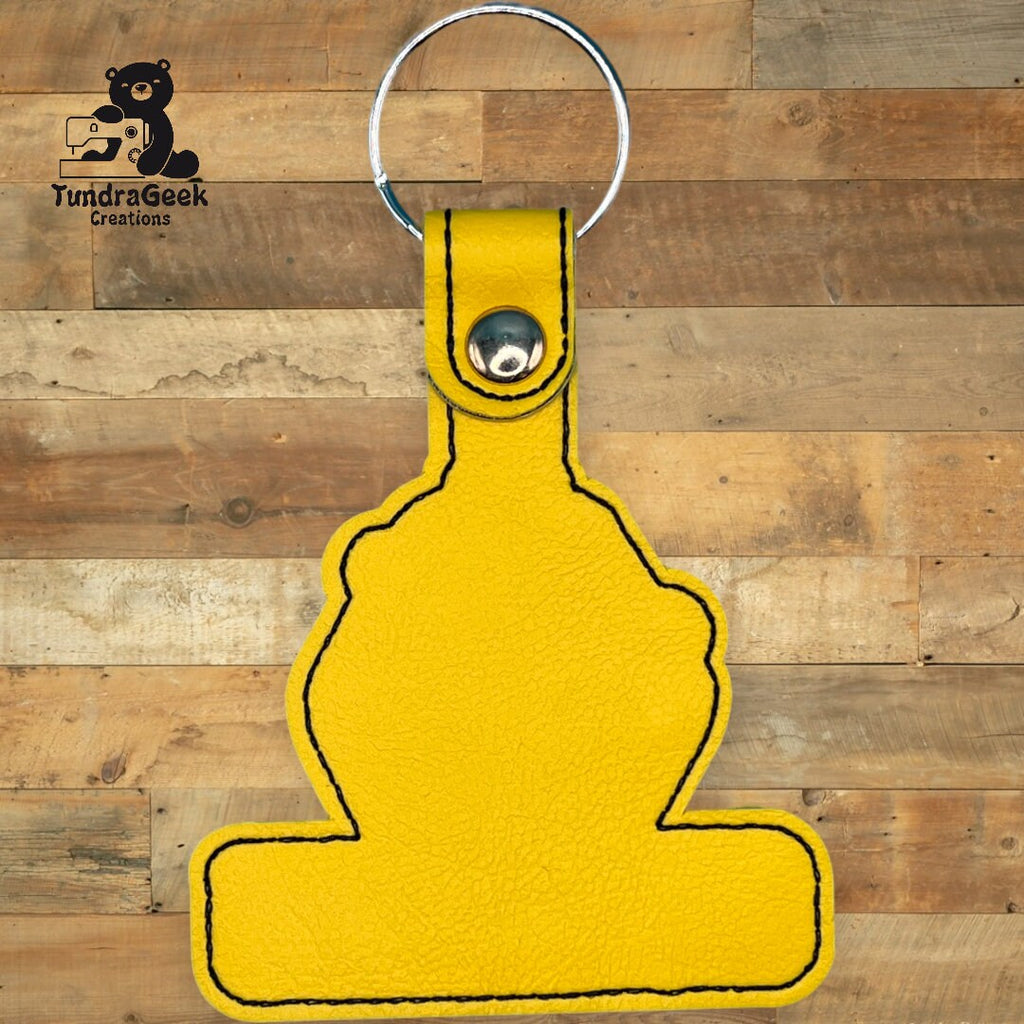 Yellow keychain with black stitching, shaped like an abstract figure, placed on a wooden background. TundraGeek Creations logo is visible in the top left corner.