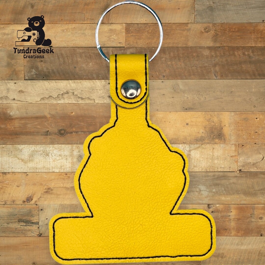 Yellow keychain with black stitching, shaped like an abstract figure, placed on a wooden background. TundraGeek Creations logo is visible in the top left corner.