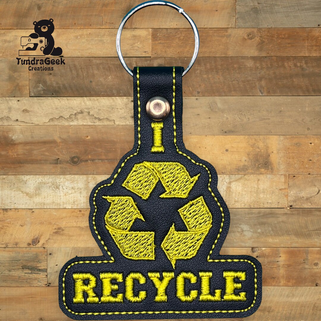 A black and yellow keychain with embroidered text and symbol reading I recycle on a wooden background.