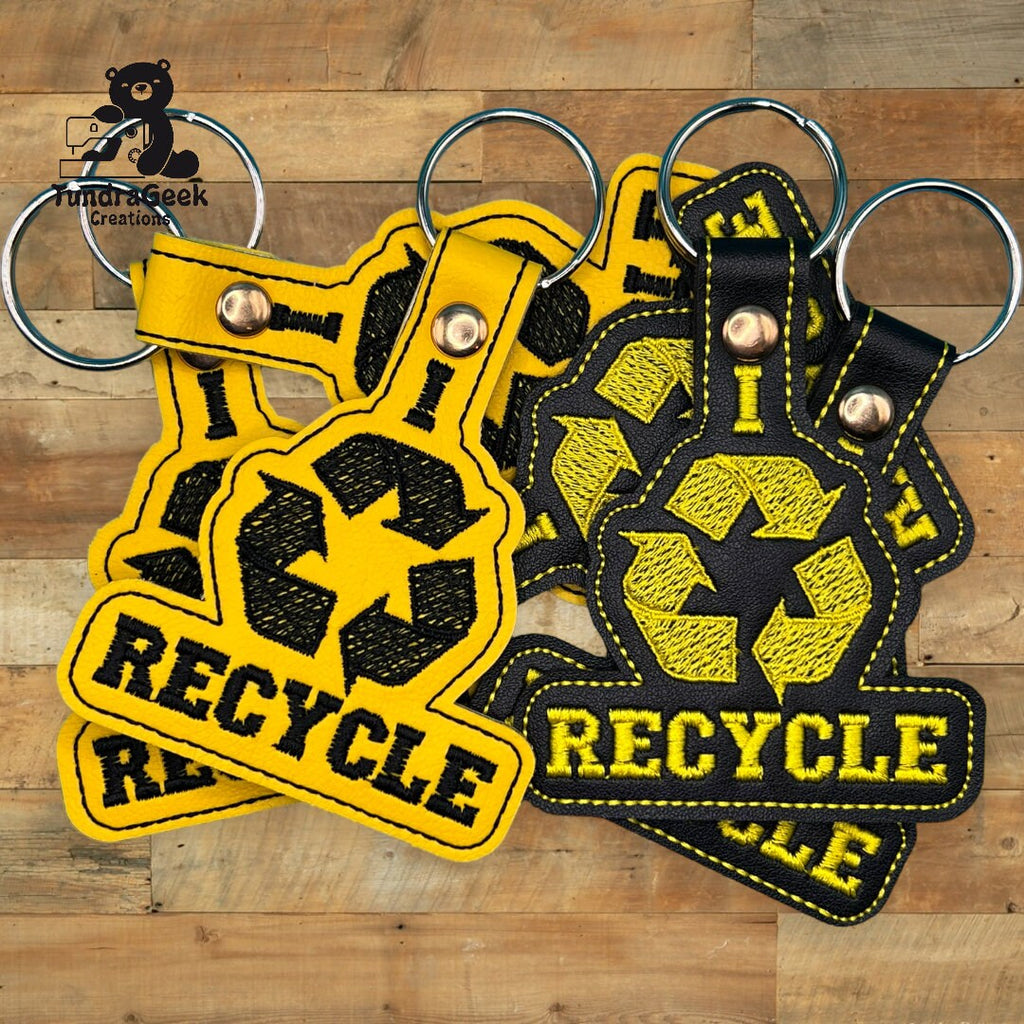 Five black and yellow keychains with the recycle symbol and the word RECYCLE are displayed on a wooden surface.