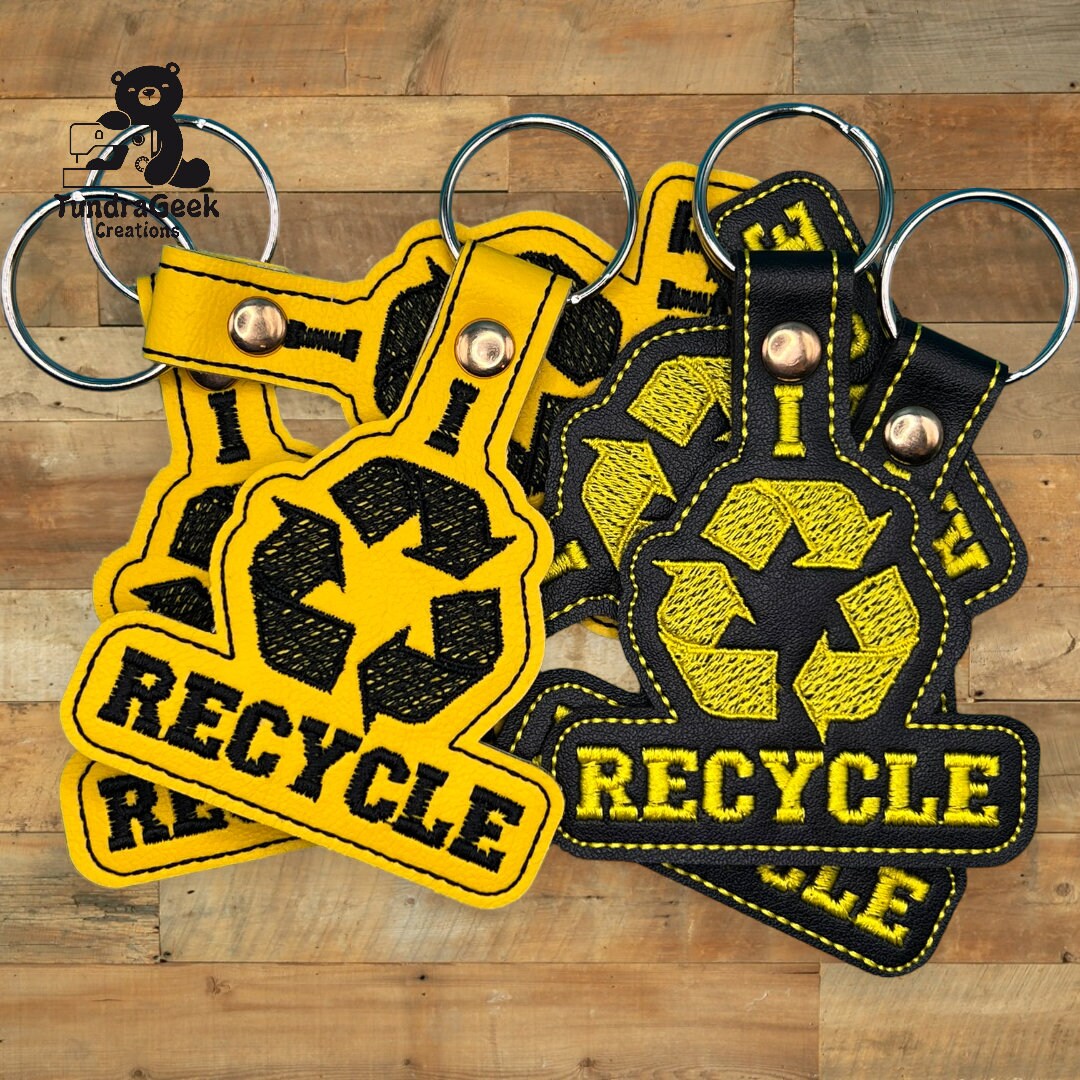 Five black and yellow keychains with the recycle symbol and the word RECYCLE are displayed on a wooden surface.