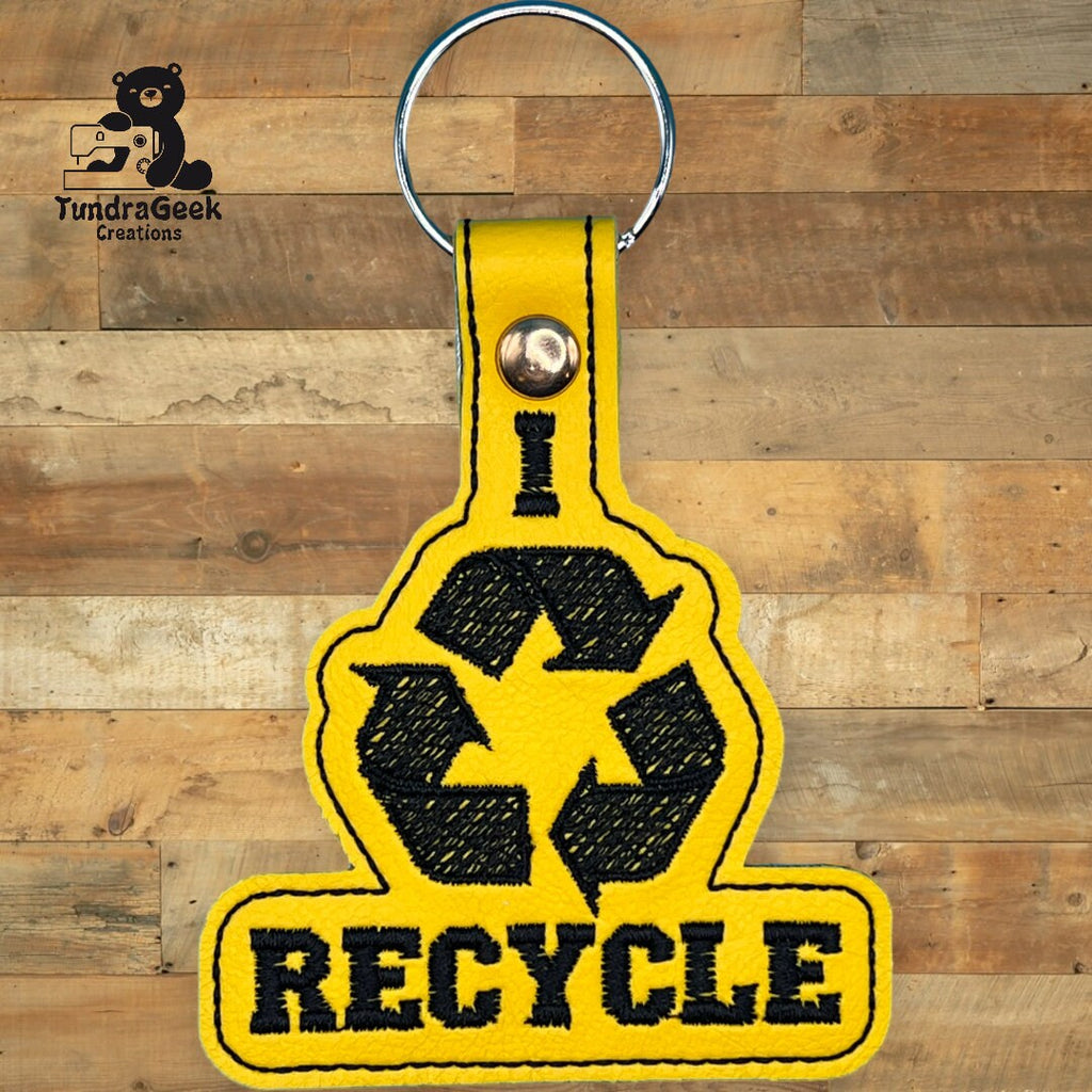 A yellow keychain with a black recycling symbol and the word RECYCLE embroidered below, attached to a silver keyring, is displayed on a wooden background.