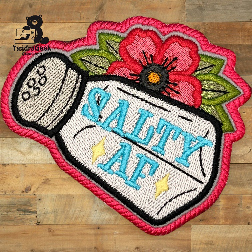 Embroidered patch of a salt shaker labeled Salty AF with a red flower and green leaves on a wooden background.