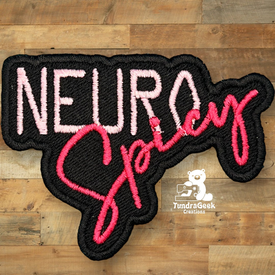 A black patch with NEURO Spicy embroidered in pink and red thread, placed on a wooden surface. The TundraGeek Creations logo is at the bottom right.