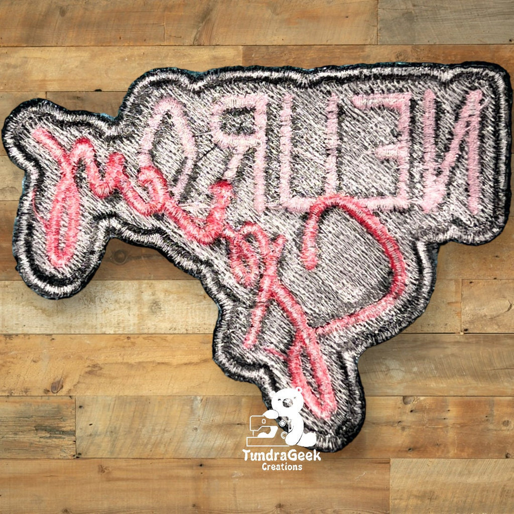 A patch shaped like Texas with pink cursive text reading Hello Y’all in reverse, displayed on a wooden background.