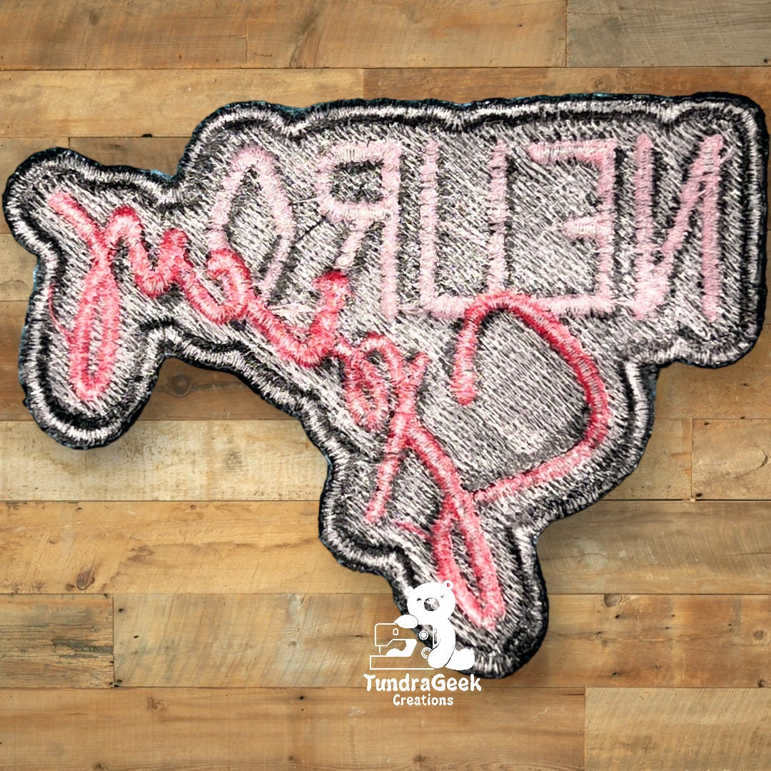 A patch shaped like Texas with pink cursive text reading Hello Y’all in reverse, displayed on a wooden background.