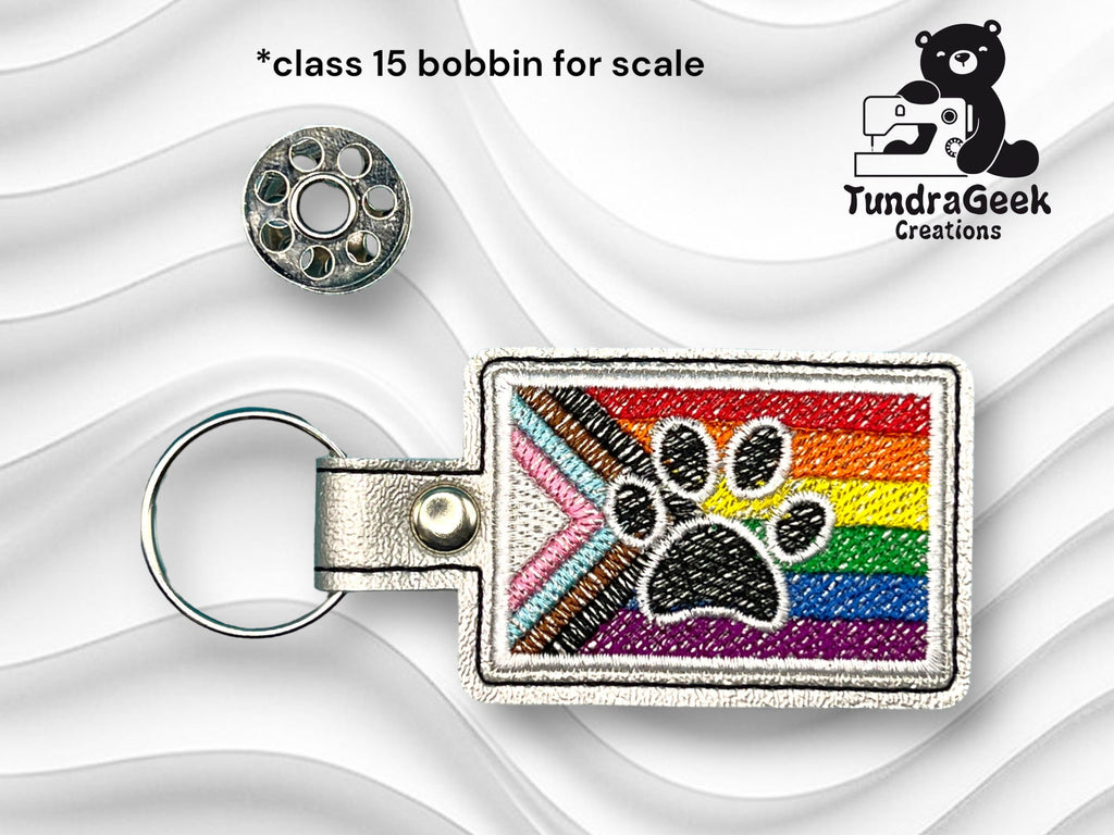 Rectangular keychain with a black paw print on a rainbow Pride flag background, next to a metal bobbin for scale. TundraGeek Creations logo in top right corner.