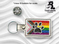 Rectangular keychain with a black paw print on a rainbow Pride flag background, next to a metal bobbin for scale. TundraGeek Creations logo in top right corner.