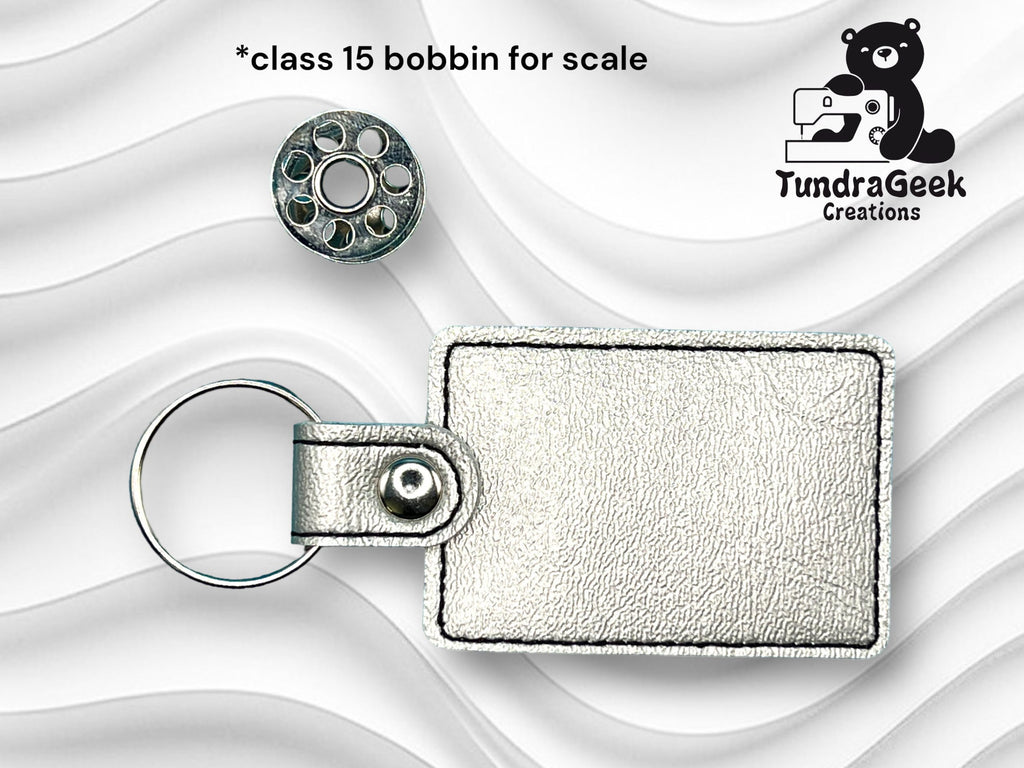 A metallic keychain with a rectangular leather tag is shown next to a class 15 sewing machine bobbin for size comparison. TundraGeek Creations logo is visible in the upper right corner.