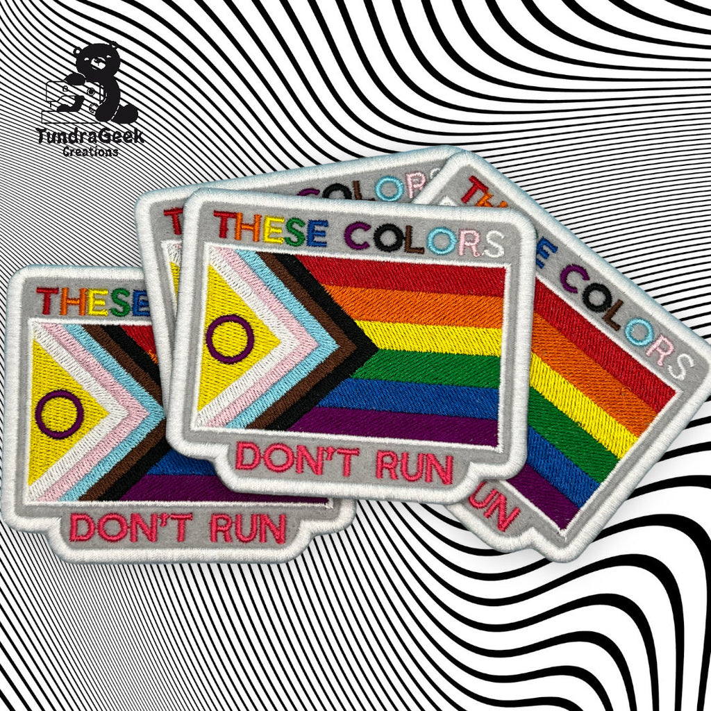 Three rectangular patches display the Progress Pride flag and the words THESE COLORS DONT RUN on a black and white wavy optical illusion background.
