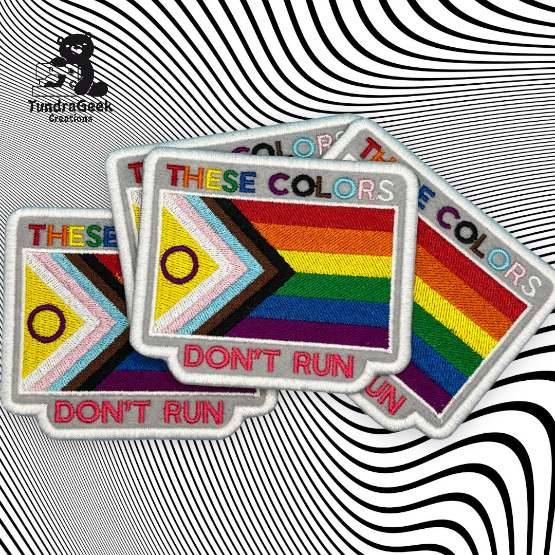 Three rectangular patches display the Progress Pride flag and the words THESE COLORS DONT RUN on a black and white wavy optical illusion background.