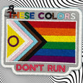 A patch with the Progress Pride Flag and the Intersex symbol, featuring the text THESE COLORS DONT RUN embroidered above and below the flag.
