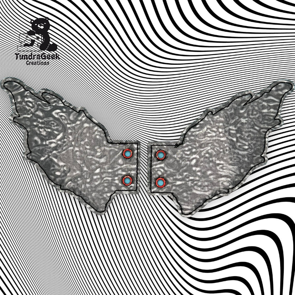Two metallic, wing-shaped objects with textured surfaces are placed against a black and white, wavy optical illusion background. The Fundi Geek Creations logo appears in the top left corner.