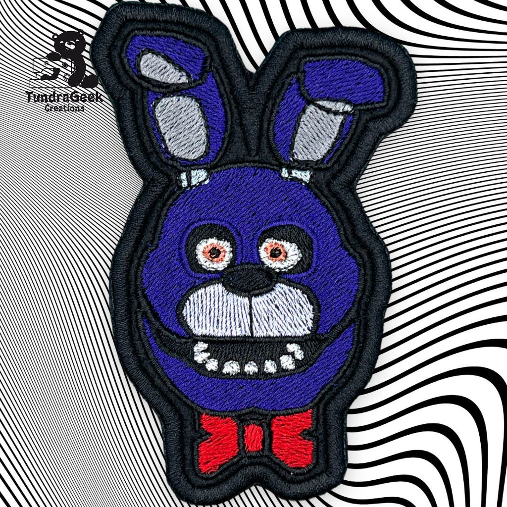 Embroidered patch of a blue cartoon rabbit face with red bow tie and wide, toothy smile, set against a black-and-white wavy line background.