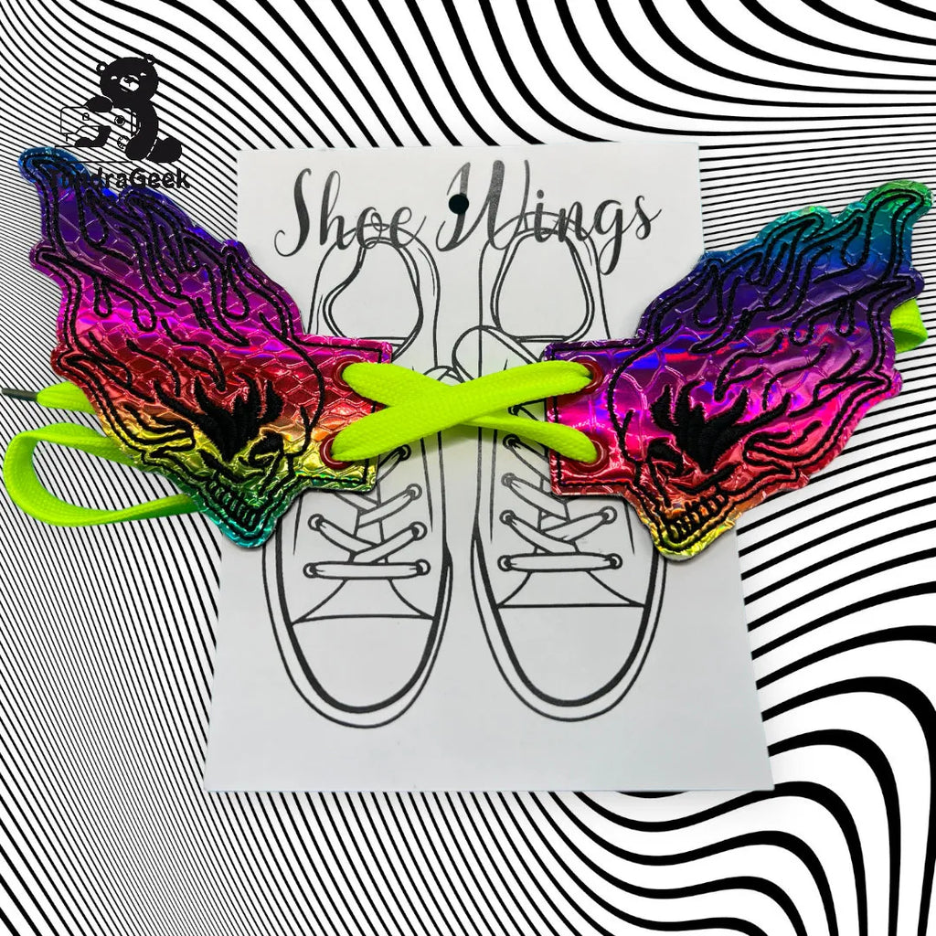 A pair of rainbow-colored, skull-shaped shoe accessories are displayed on a card with a sneaker illustration and lime green laces, set against a black-and-white swirling pattern background.