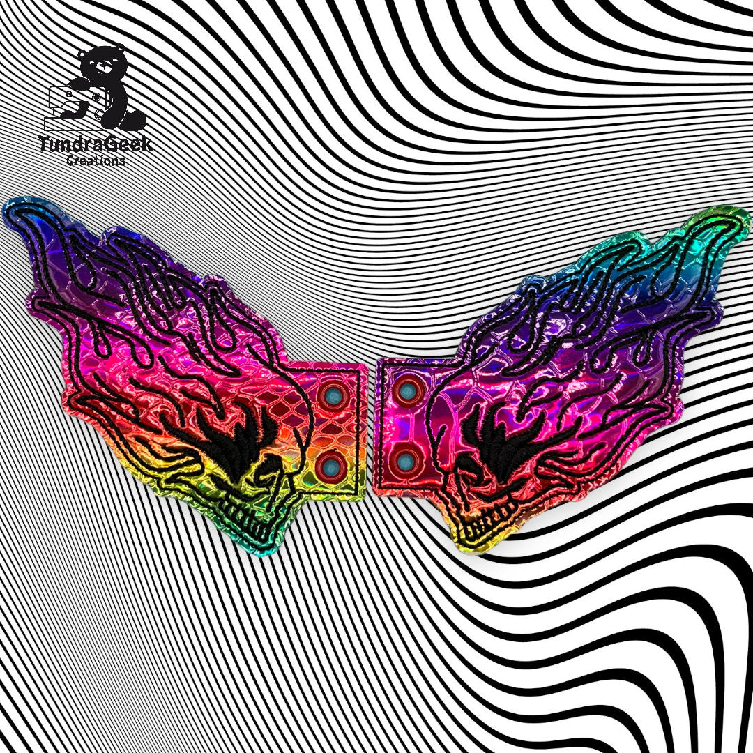 A pair of rainbow-colored, wing-shaped belt buckles on a black and white wavy striped background with the FundrGeek Creations logo in the corner.