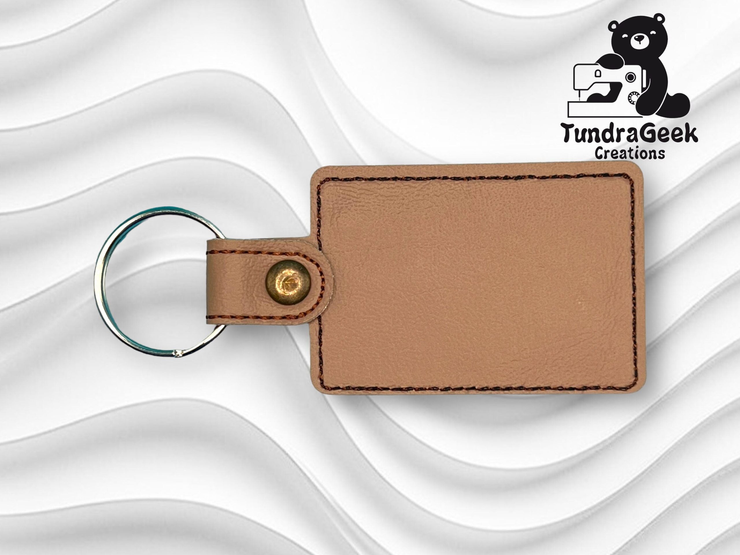Rectangular brown leather keychain with black stitching and gold snap, displayed on a wavy white background with the TundraGeek Creations logo in the top right corner.