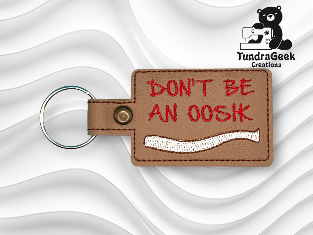 A brown leather keychain with red text reading DONT BE AN OOSIK and a white embroidered shape, set against a wavy white background. TundraGeek Creations logo is in the top right corner.