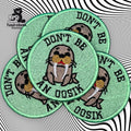 Five green embroidered patches featuring a cartoon walrus and the text DONT BE AN OOSIK, arranged in an overlapping pattern on a black and white wavy background.