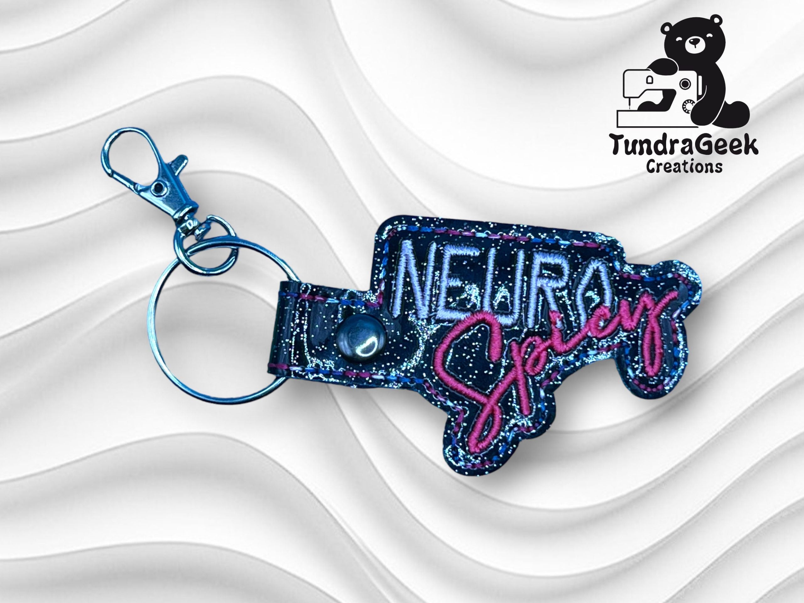 A glittery keychain reading NEURO Spicy in blue and pink, with a metal clasp and ring, displayed on a wavy white background. TundraGeek Creations logo is in the top right corner.