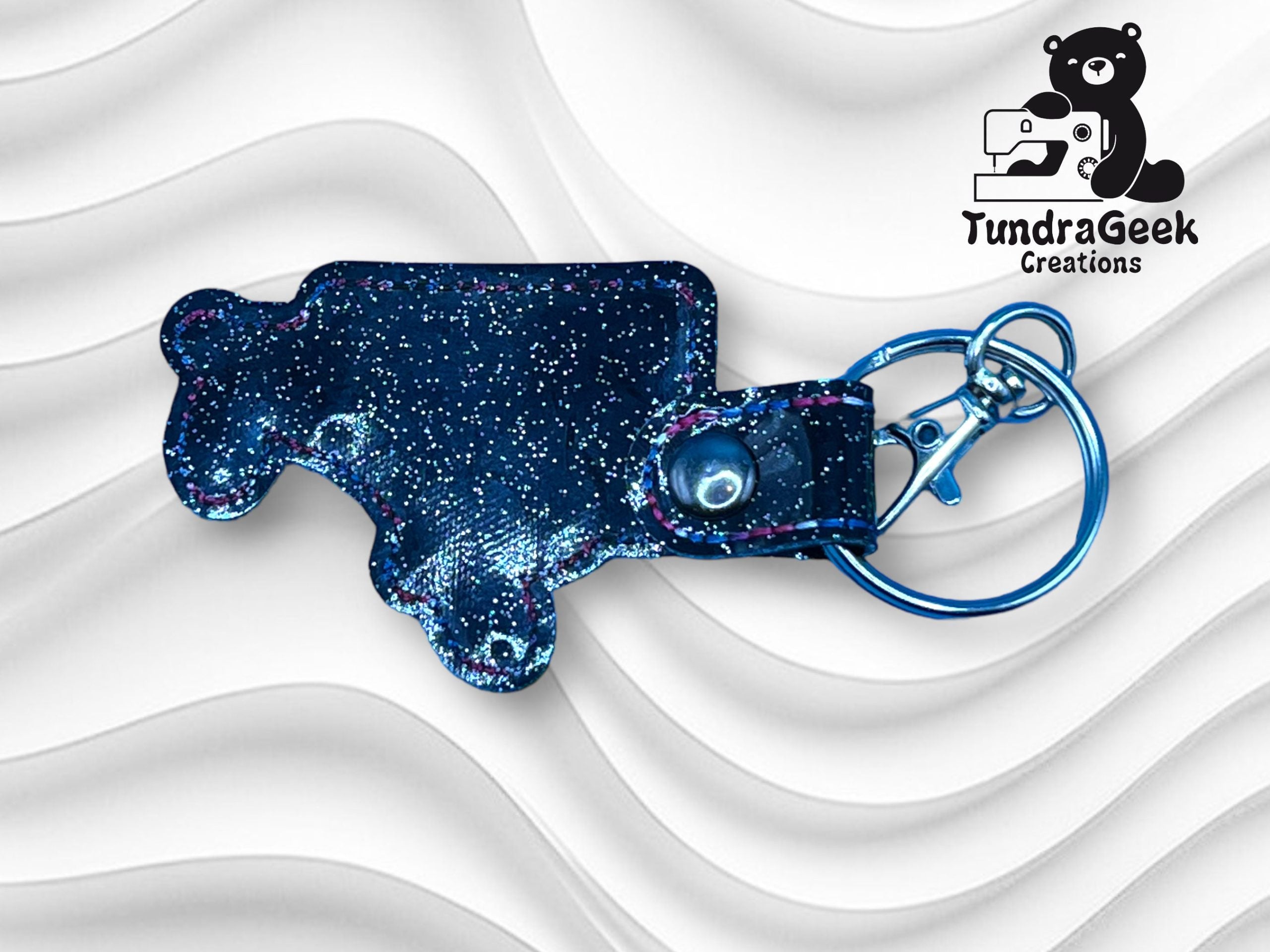 A glittery blue bear-shaped keychain with a snap button and metal keyring, displayed on a wavy white background with the TundraGeek Creations logo in the corner.