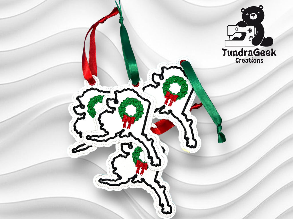 Three Alaska-shaped ornaments with holiday wreaths and green ribbons are displayed on a white textured background. The TundraGeek Creations logo appears in the upper right corner.