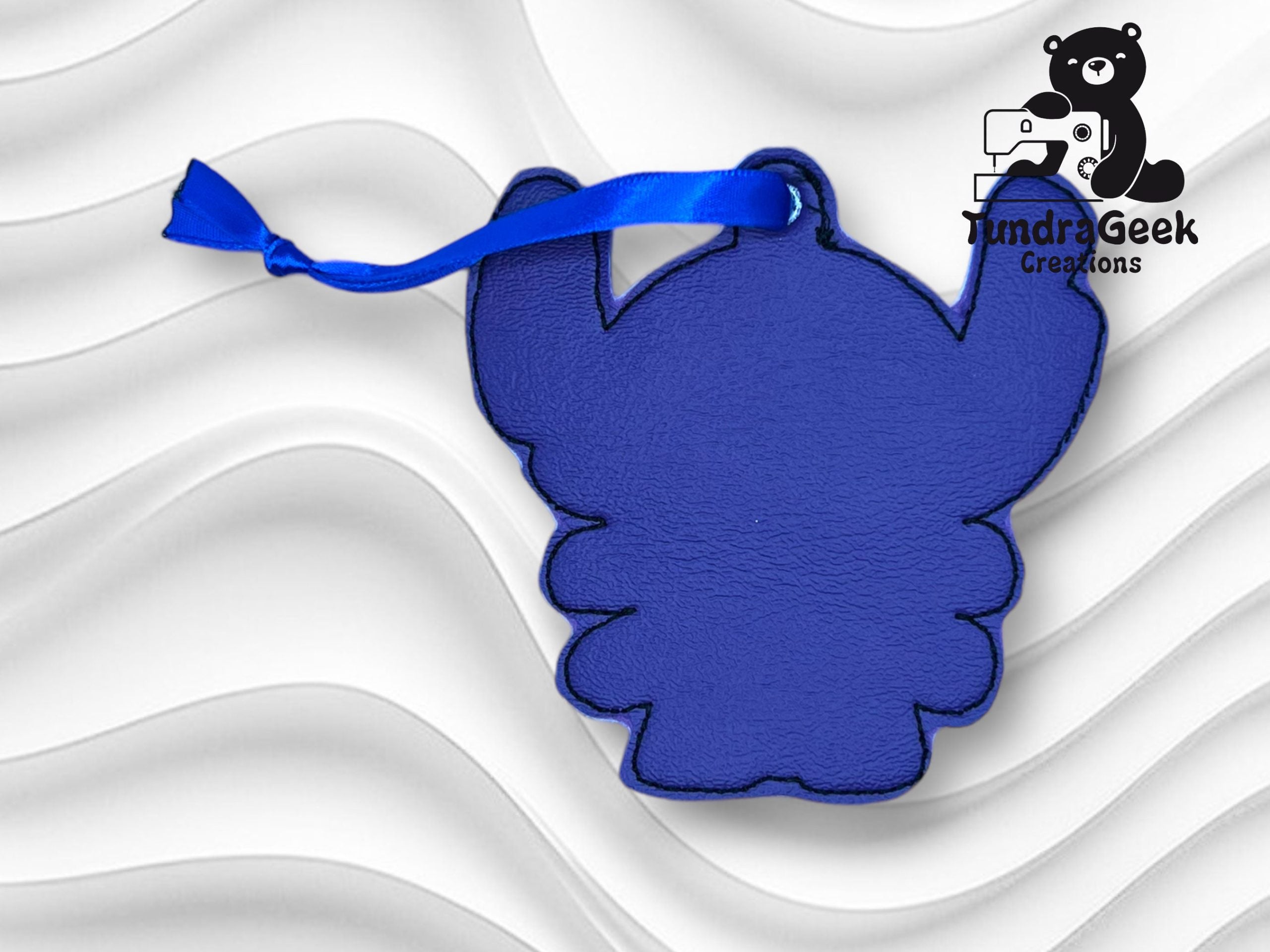 Blue faux leather keychain with a ribbon, shaped like a cartoon figure, displayed on a wavy white background with the Tundra Geek Creations logo in the corner.