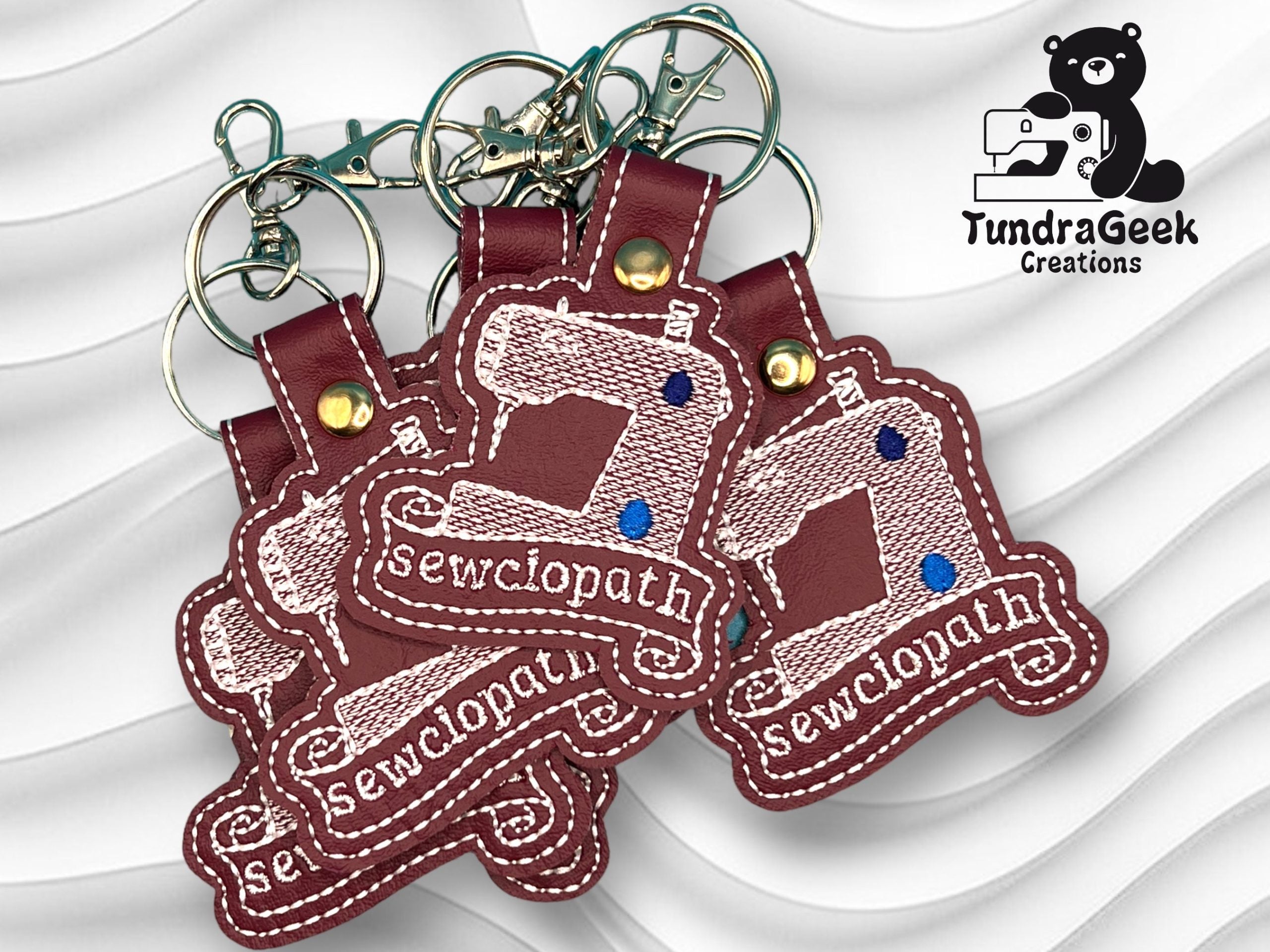 Four maroon keychains shaped like sewing machines with Sewciopath embroidered on each, displayed on a white textured background with a TundraGeek Creations bear logo.