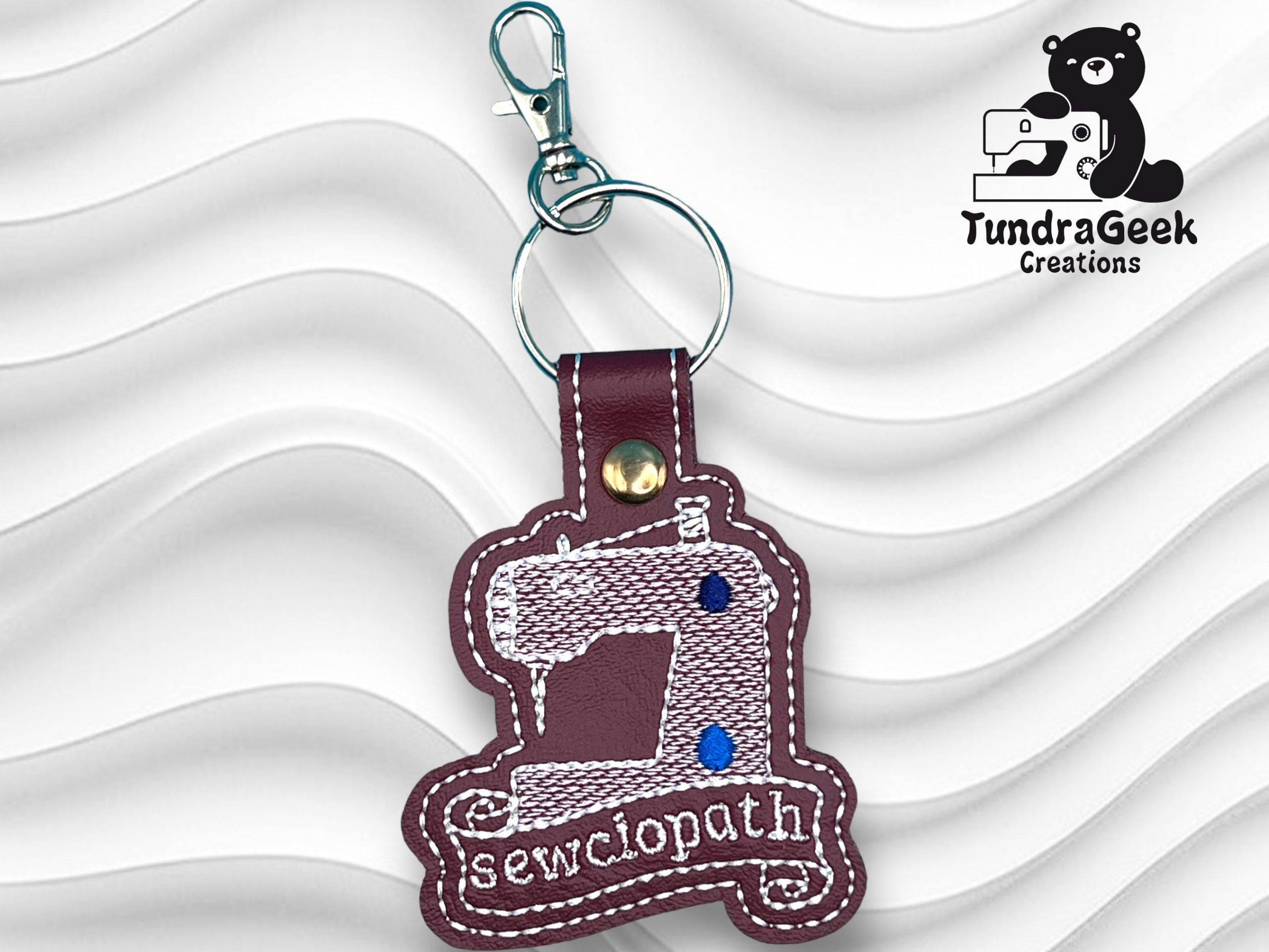 Keychain shaped like a sewing machine with the word sewciopath embroidered on it, displayed on a textured white background; TundraGeek Creations logo in the top right corner.